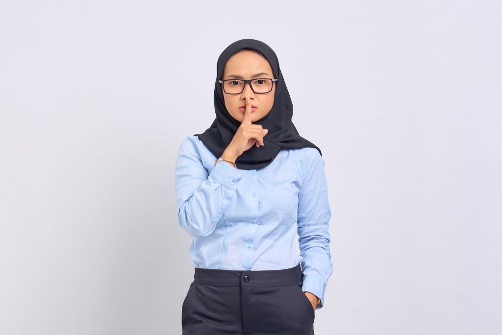 Asian Muslim woman in hijab making a quiet gesture with a serious expression, wearing eyeglasses against a white background.