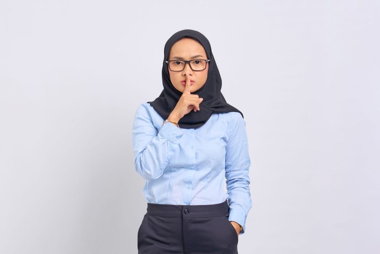 Asian Muslim woman in hijab making a quiet gesture with a serious expression, wearing eyeglasses against a white background.
